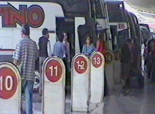 Travelers waiting to board buses at a bus station, where several buses are parked in numbered gates