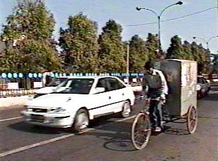 A white car driving next to a man riding a bicycle on the street, pulling an item on a cart