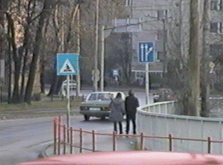 Two women walking on a sidewalk next to a street, where a sedan is driving
