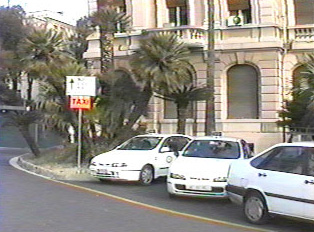 Three white taxis parked next to an orange taxi sign