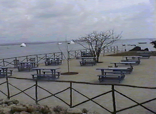 A pier by the ocean, empty of people, with several tables and benches, and boats on the water in the background