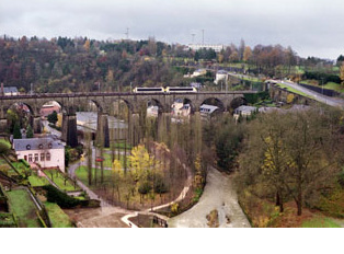 A train runs on a stone bridge above houses and a green landscape