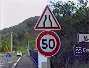 A red and white road sign indicating the road number and the merging of two lanes